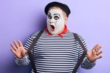 close up portrait of an amazed clown in striped sweater with open mouth stretching his suspenders and looking at the camera,