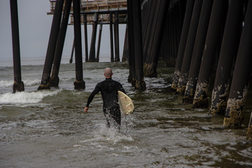Surfs up at the pier. Lone surfer running between the pilons of the pier, carrying his surfboard at his side, rushing to catch the waves.