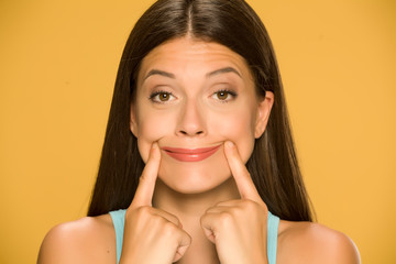 Young woman forcing her smile with her fingers on yellow background