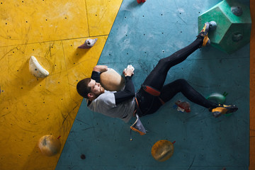 Side view of motivated active physically challenged boulderer with beard hanging at colourful climbing wall, holding large artificial rock, trying to fix his leg at feet hold. Indoor sport concept. - Powered by Adobe