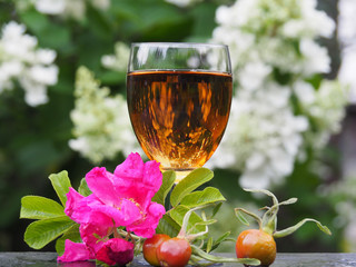 Useful tincture in a wine glass, a flower with leaves of wild roses, rose hips in the garden on a background of white flowers in the summer. Bright and beautiful picture for decoration and design. 