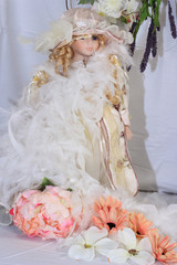 Old porcelain doll with golden curls, white background, light pink gown with white lace, with a white feather boa. Peach silk flowers laying at her feet. Green & purple foliage hanguibg behind her.