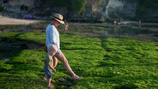 Slow motion of young caucasian mother and two-years old boy are walking on green algae at low tide. Balangan beach, Bali, Indonesia