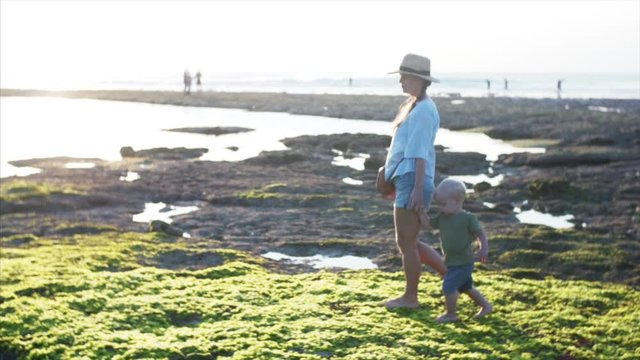 Slow motion of young caucasian mother and two-years old boy are walking on green algae at low tide enjoying evening sun. Balangan beach, Bali, Indonesia