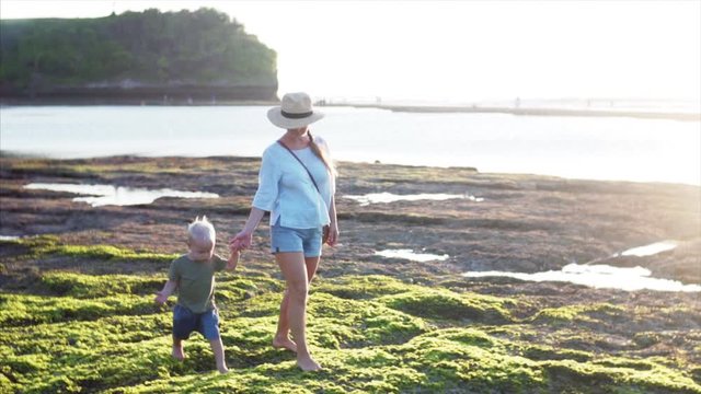 Slow motion of young caucasian mother and two-years old boy are walking on picturesque green algae at low tide enjoying evening sun. Balangan beach, Bali island, Indonesia