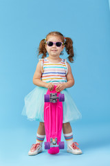 Stylish little toddler girl child in glasses with skateboard over blue background