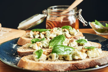 Close up on a plate of toasts with roquefort cheese, walnuts, honey and basil. A jar of honey in the background 