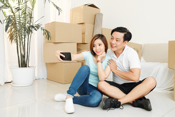 Cheerful Vietnamese couple taking photo on smartphone after moving in new apartment or house