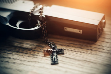 On a wooden table lies a gun lit by sunlight and black carved Holy cross with black beads on the chain.