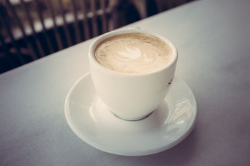 Cup of coffee with milk on marble table with wooden chairs in unfocused background, vintage style