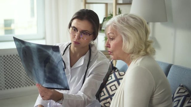 Confused aged woman speaking with practitioner wearing in white medical coat. They sitting on soft couch or sofa in modern interior apartment, checking problems and discuss x-ray picture of lung