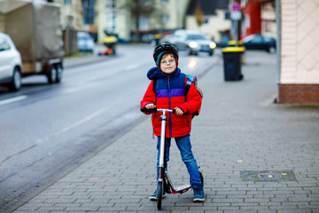 Gorgeous little school kid boy riding on scooter on way to elementary school. Child with safety helmet, school bag on rainy autumn or winter cold morning. Traffic in the city and schoolchildren