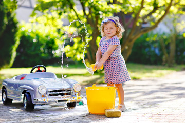 Cute gorgeous toddler girl washing big old toy car in summer garden, outdoors. Happy healthy little...