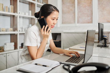 Young Asian receptionist checking schedule on laptop screen when answering phone call on client