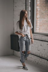 Seductive portrait of young sexy girl with brunette hair in shirt, jeans and sneakers on white background in room near window