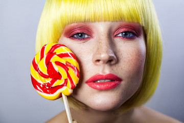 Beauty portrait of cute young female model with freckles, red makeup and yellow wig, holding...