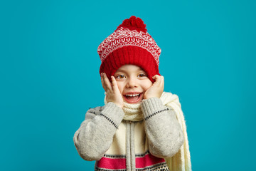 Funny girl in winter hat, put her hands to cheeks, cute smile, expresses a cheerful mood and surprise, children's emotional portrait on blue background.