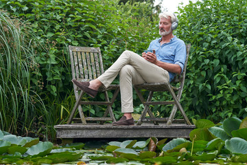 Mature Man Relaxing In Garden Listening To Music On Wireless Headphones On Jetty By Lake