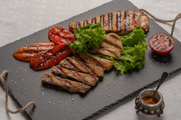 Grilled turkey breast sliced steaks with baked peppers,lettuce and tomato on a black board. Next to the pepper shaker with red pepper. Horizontal orientation, close-up