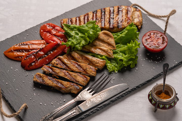 Grilled turkey breast steaks with baked peppers,lettuce and tomato on a black board with knife and fork. Next to the pepper shaker with red pepper. Horizontal orientation, close-up
