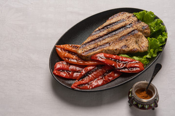 Grilled Turkey breast steak with baked peppers and lettuce leaves in a black plate on a light linen background. Next to the pepper shaker with red pepper. Horizontal orientation, copy space