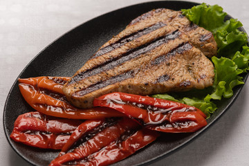 Grilled turkey breast steak with baked peppers and lettuce leaves in a black plate on a light linen background. Close-up, top view