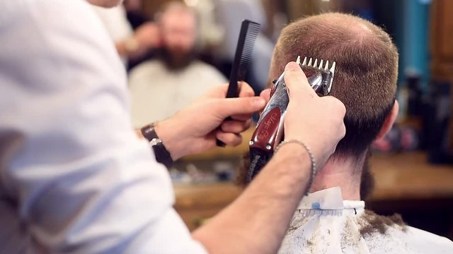 Male client sitting back to camera in cape while barber hands working with electric razor and comb. Making stylish haircut in barbershop. Blurred reflection in mirror on background. Cropped filming