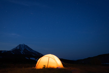 Night photo of the camping site in Kamchatka, Russia. Yellow tent in the darkness. Stars in the sky. Silhouette of smmetrical cone of Vilyuchik volcano in dark blue  background.
