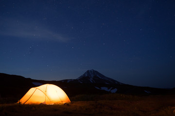Fototapeta na wymiar Night photo of the camping site in Kamchatka, Russia. Yellow tent in the darkness. Stars in the sky. Silhouette of smmetrical cone of Vilyuchik volcano in dark blue background.