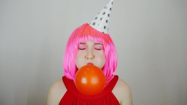 A woman in a pink wig inflates a red ball. Birthday celebration.
