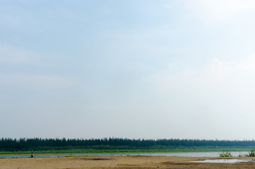 Obraz premium Girl traveler resting on the edge of the old pier on the banks of the river vilyu with spruce forests and the tundra of Yakutia in the Far North of Russia looking at the phone on a warm day.
