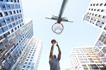 Low angle action shot of African basketball player shooting slam dunk against sky in urban...