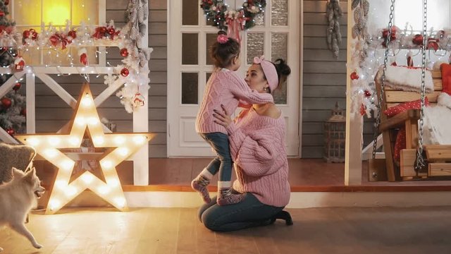 Happy mother having fun with her kid daughter at Christmas porch. Mother tickling her little daughter in slow motion. Beautiful mother and daughter in pink pullovers at Christmas day. Full length shot