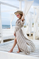 Small (8 years old) pretty cheerful girl in a white striped dress and in pink sunglasses stands on a wooden white pier.8 years old girl resting on the tropical beach alone on a warm sunny day.Child po