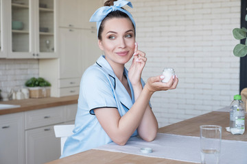 Beautiful fit brunette woman at home in pajama. She do beauty procedures - apply face cream. Natural light. Lifestyle healthcare concept