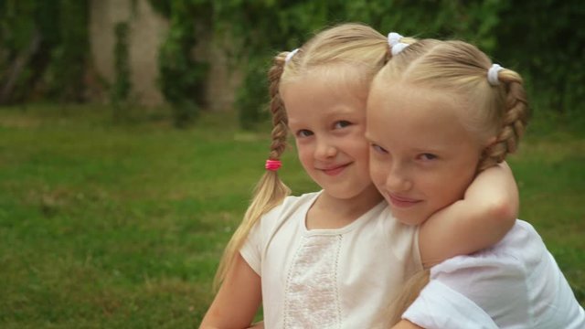 Two smiling sisters girls hug tightly each other in a backyard in sunny day. Two blonde sisters spend time together. Concept of family and happy childhood