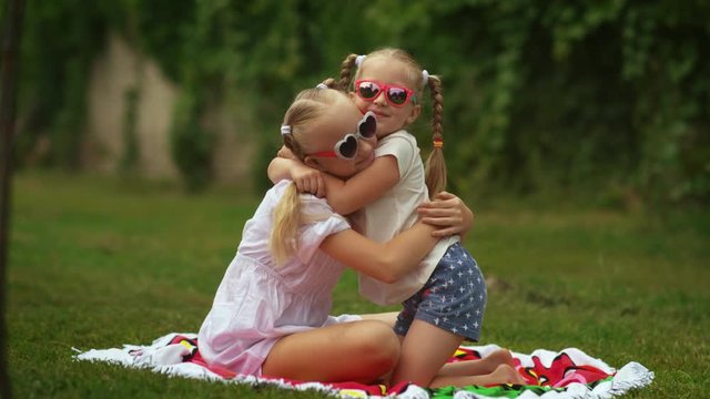 Two children sisters in sunglasses hug tightly each other in a backyard in sunny day. Two blonde sisters spend time together. Concept of family and happy childhood