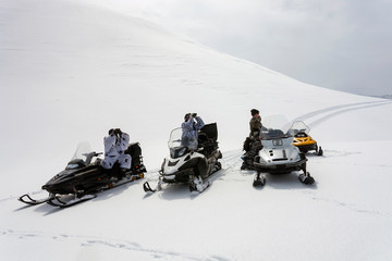 Hunters in winter camouflage on snowmobiles are looking through binoculars.
