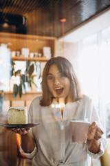 portrait of attractive and positive girl dressed in stylish clothes holds a mug of coffee and a plate with a large piece of cake and looks appetizing at them
