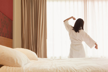 Fototapeta na wymiar Woman stretching in bed after waking up, back view. Woman sitting near the big white window while stretching on bed after waking up with sunrise at morning, back view.