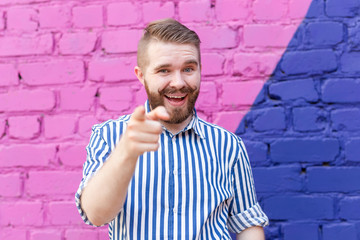 Funny young stylish guy with a mustache and a beard in a stylish shirt posing on of a blue-purple brick wall and pointing fingers into the camera. Concept of promotions and discounts.