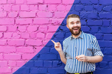 Portrait is a nice young student guy with a mustache and a beard posing against a blue-violet brick wall showing a finger on something useful. Place for your advertising.