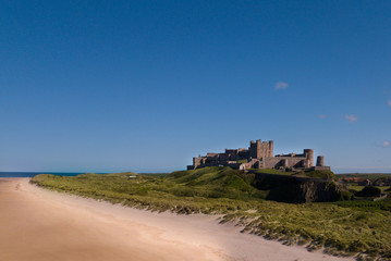 Fototapeta premium Aerial overhead view of Bamburgh Castle in the beautiful quant old Northumbrian village. Drone shot view across the sand dunes and beach set against a brilliant blue sky.
