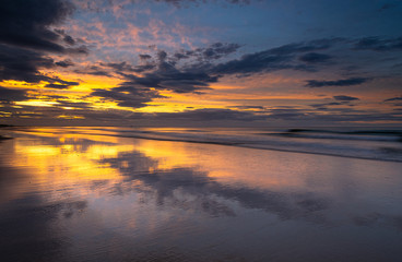 Fototapeta premium Coastline beach sunset on the shore at Bamburgh, Northumberland in north east England. Incoming tide on the sand providing reflections and symmetry to the colourful and dramatic skies at dusk.