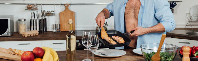 panoramic shot of man holding frying pan and cooking fish in kitchen
