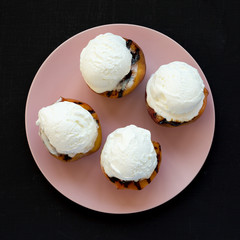 Homemade grilled peaches with vanilla ice cream on a pink plate on a black surface, top view. Flat lay, from above, overhead.