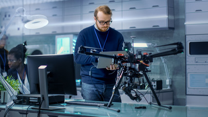 Young Male Engineer Programs Drone while Holding Laptop in His Hands. He Works in a Bright Modern High-Tech Laboratory.