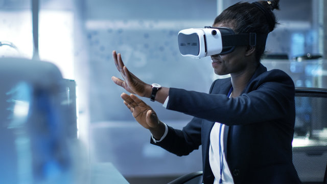 African-American Female Virtual Reality Engineer/ Developer  Wearing VR Headset Creates Content. She's Alone in a Modern Laboratory/ Research Center.