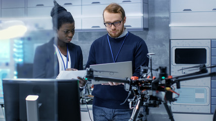 Caucasian Male and Black Female Engineers Working on a Drone Project with Help of Laptop and Taking...