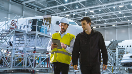 Fototapeta premium Team of Aircraft Maintenance Mechanics Moving through Hangar. Holding Tablet Computer